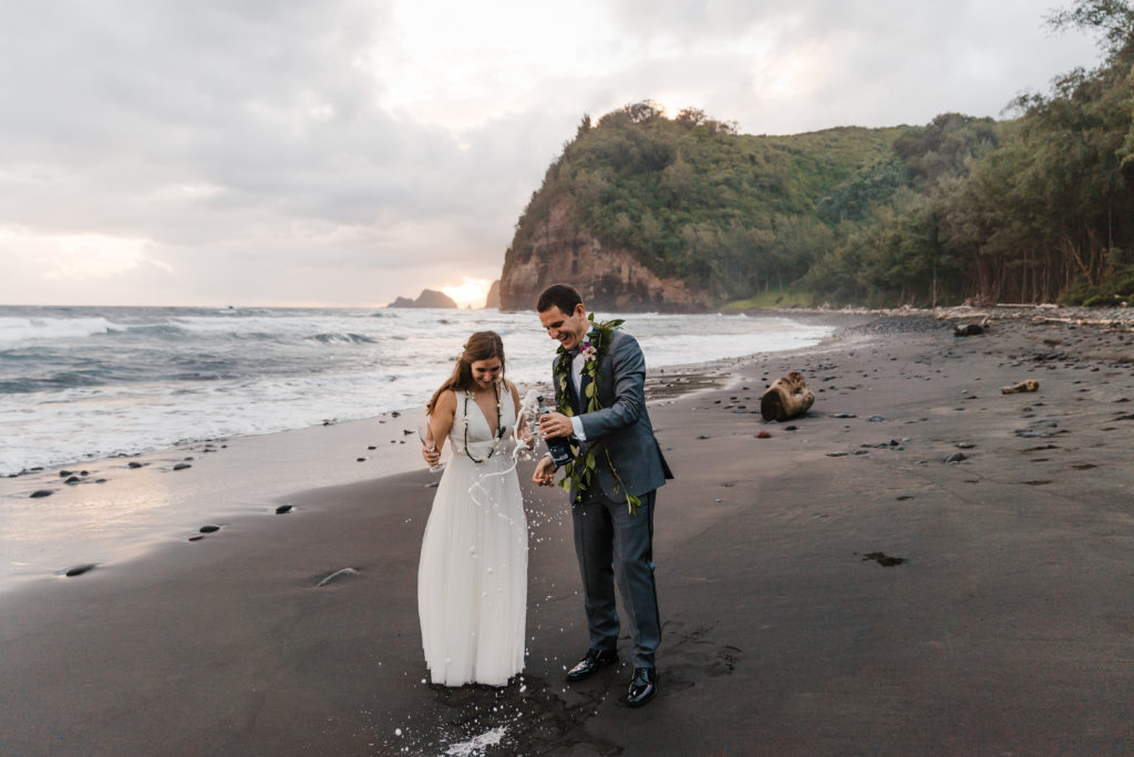 bride and groom popping champagne on black sand beach in hawaii
